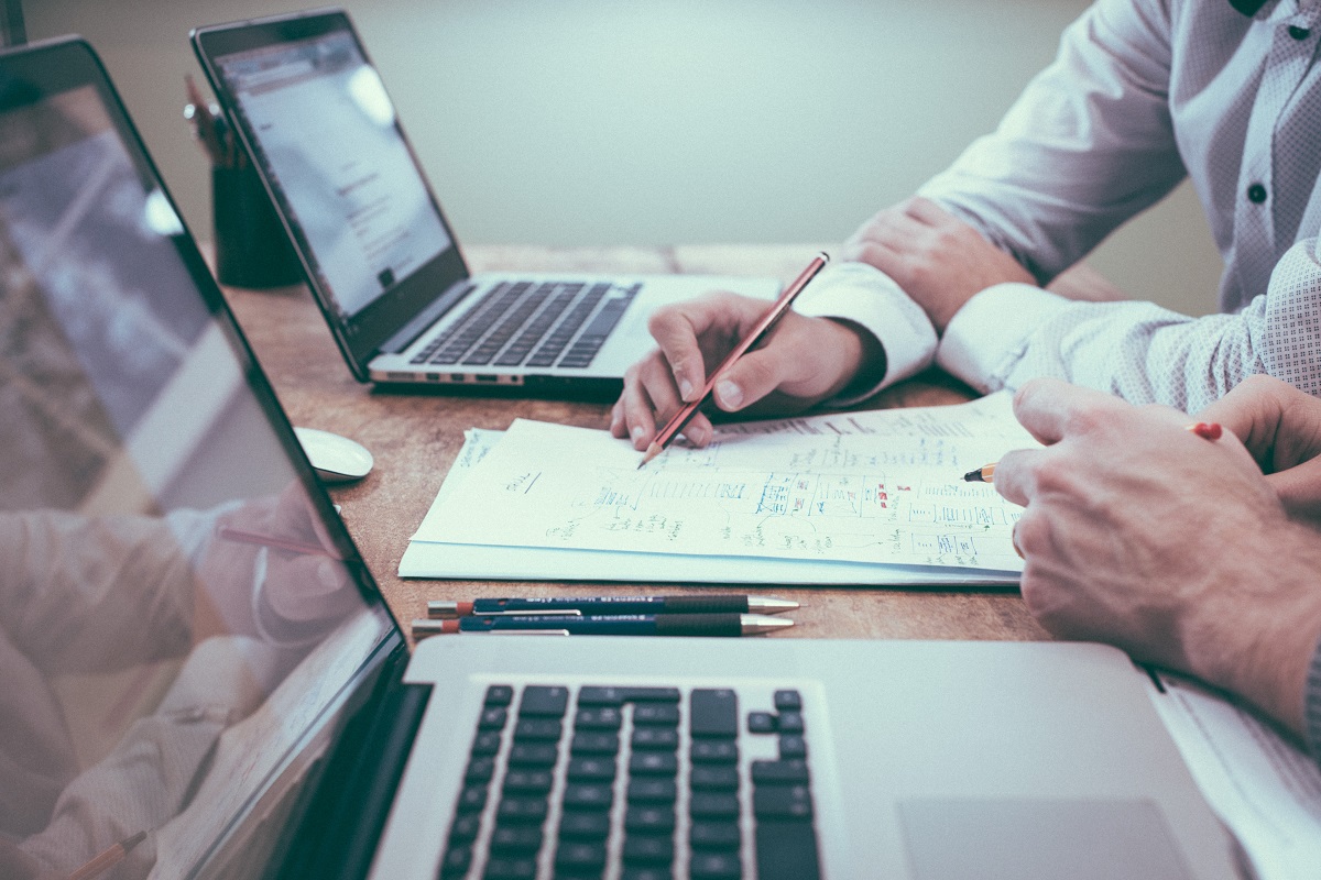 Duas pessoas trabalhando lado a lado em escritório usando notebooks e vendo dados em folhas de papel entre eles como Sales analyst