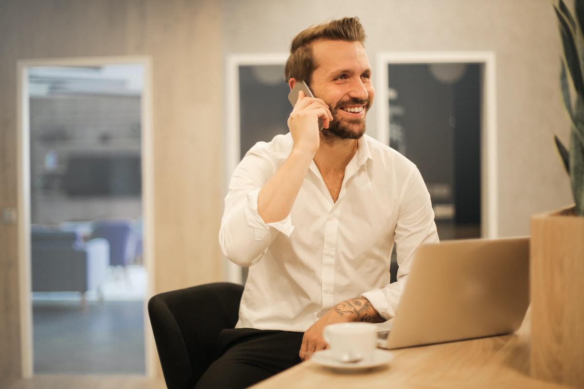 Homem conversando em celular durante ligação diante de mesa de madeira usando notebook com xícara de café branca sobre pires de mesma cor em home office