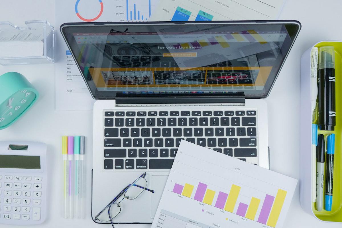 Escritório: Sistema de gestão Notebook sobre mesa branca com gráficos impressos em cima de seu teclado e óculos de grau com armação dobrada, além de itens de escritório, calculadora e relógio ao redor do mesmo