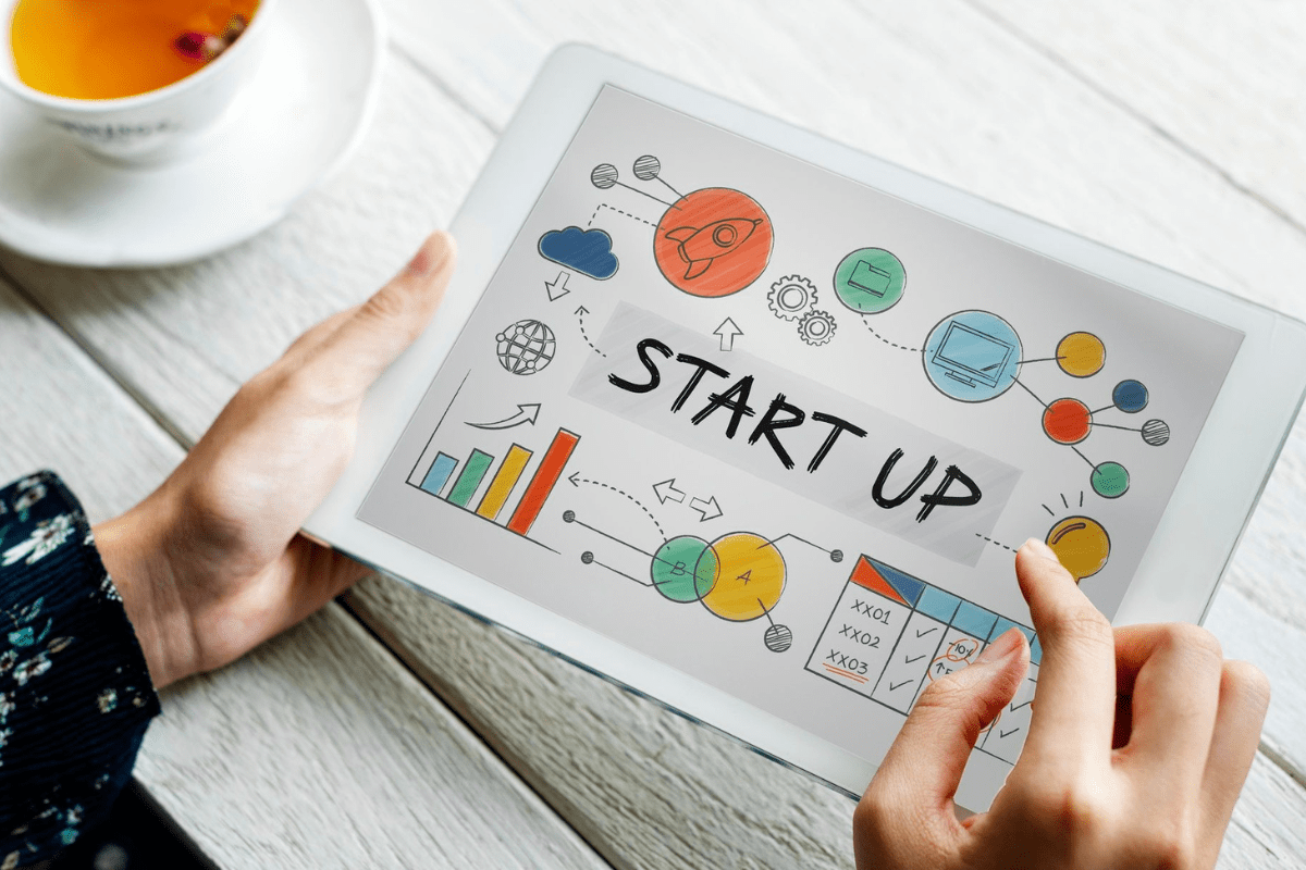 Pessoa com tablet na mão trabalhando em montagem com termo Startup e desenhos sobre o tema ao redor do mesmo