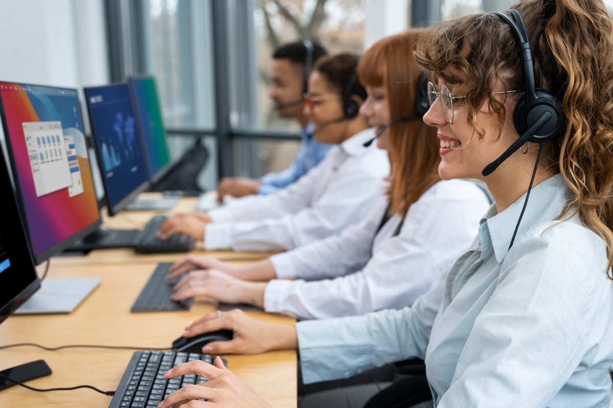 Empresa: SugarCRM Time de atendentes sentados lado a lado com roupas sociais em empresa de telemarketing diante de seus computadores e com fones headset sorridentes aplicando técnicas de SugarCRM