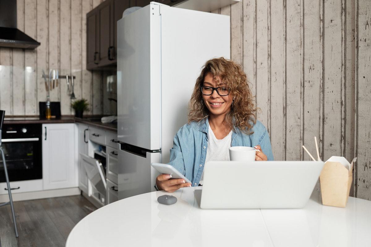 Mulher trabalhando em home office usando notebook branco sobre mesa redonda de mesma cor e usando smartphone também branco com móveis e eletrodomésticos de cozinha ao fundo