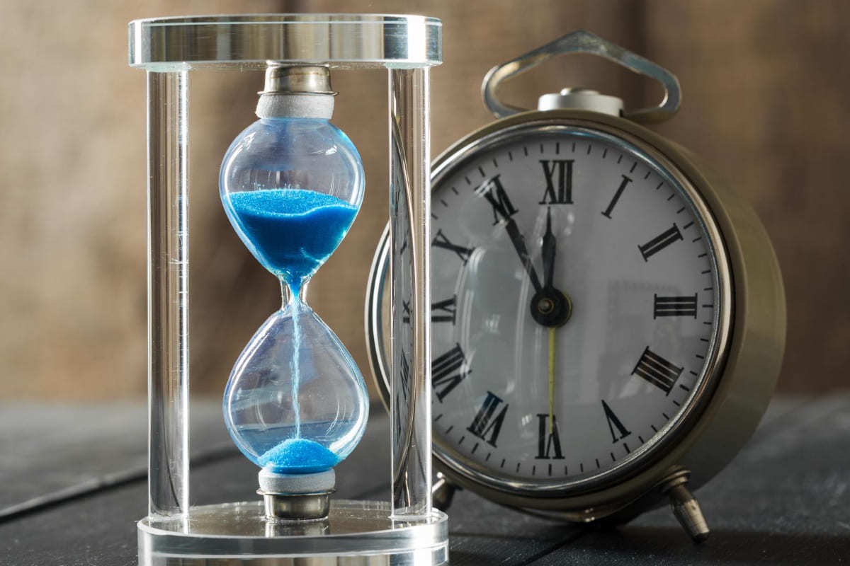 Relógio: Teste A/B para e-commerce Ampulheta com areia azul ao lado de relógio antigo com função despertador sobre mesa de madeira cinzenta
