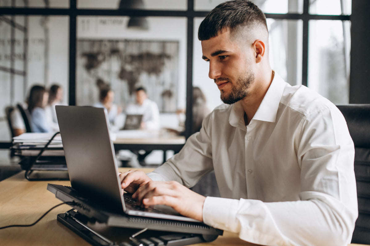 Notebook: Twitter analytics Pessoa trabalhando em notebook sobre suporte em cima de mesa de madeira usando Twitter analytics com equipe trabalhando ao fundo reunidos