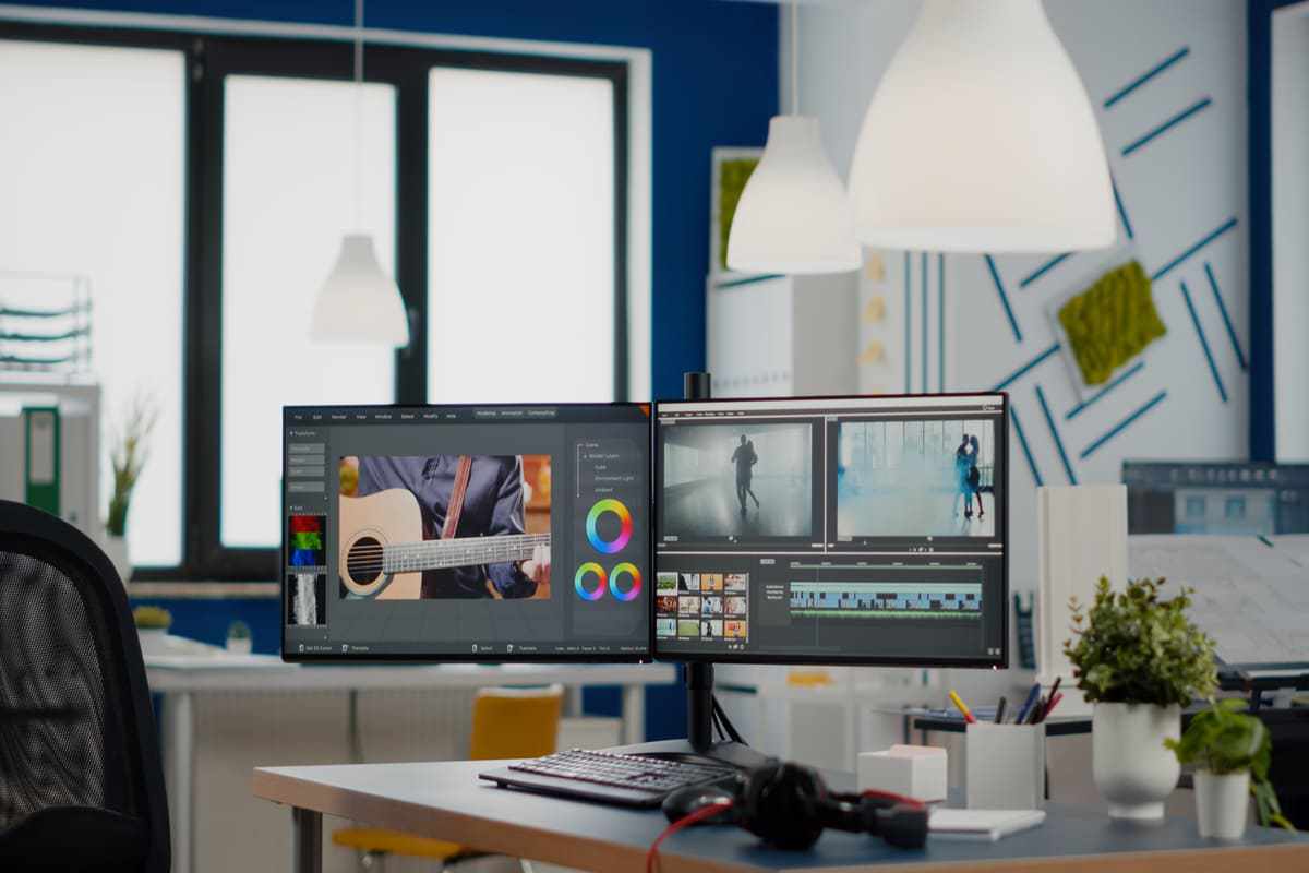 Computador sobre mesa de madeira com dois monitores com programa de edição aberto na tela do mesmo em escritório com janela de vidro de edifício ao fundo