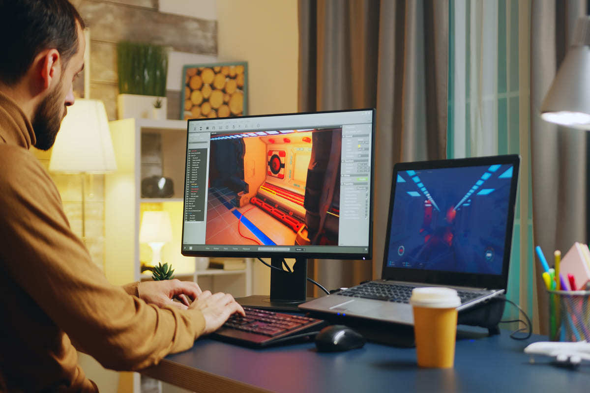 Homem a Usar Photoshop com notebook e monitor à sua esquerda sobre mesa preta com copo de café à sua direita e itens de escritório em Home office