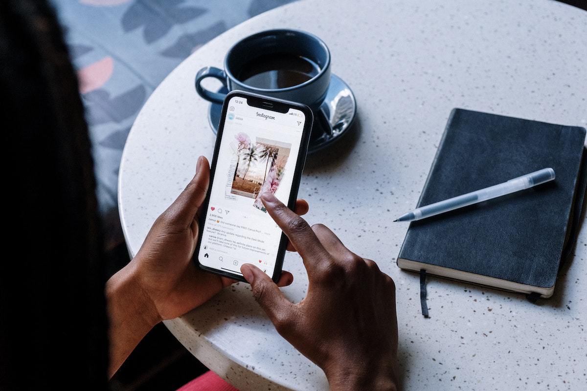 Pessoa usando celular e visualizando algo no Instagram sentada diante de mesa redonda de pedra fina com agenda e caneca e xicara de café sobre a mesma