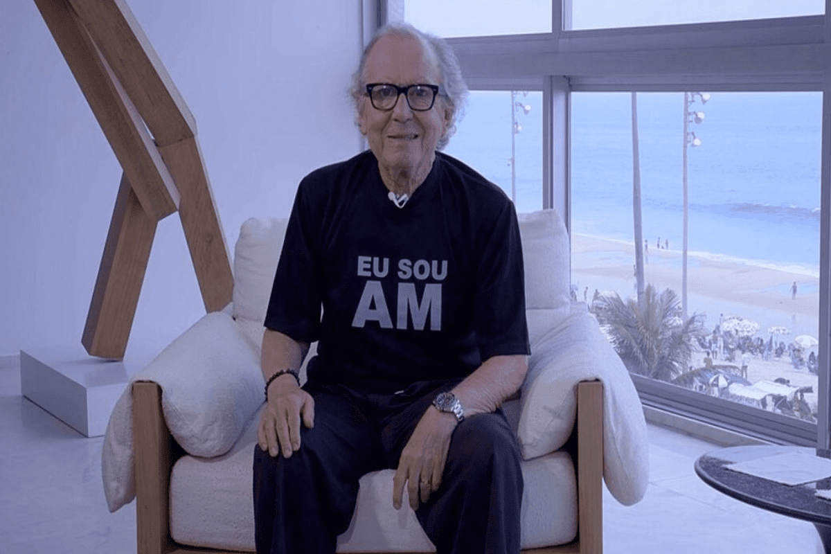 Washington Olivetto sentado em poltrona diante de janela de vidro de edifício na frente da praia e usando camisa com dizeres "Eu sou AM" em foto casual
