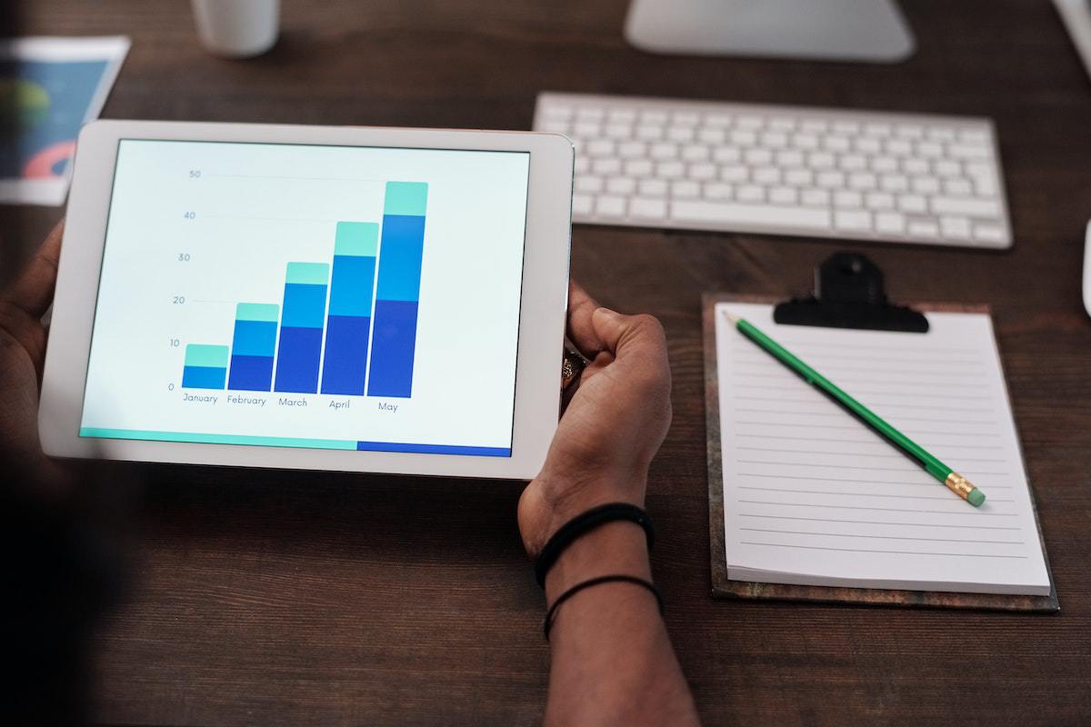 Pessoa segurando tablet com gráfico na tela ao lado de teclado branco de computador e prancheta com folhas brancas de papel e lápis sobre o mesmo