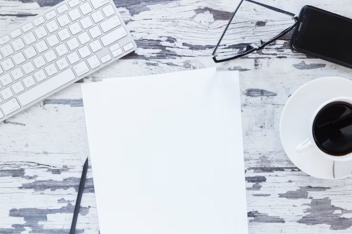 Folha: White Paper Folha branca sobre mesa branca com detalhes de tinta descascada com xícara ao lado da mesma e teclado em frente às folhas representando White Paper