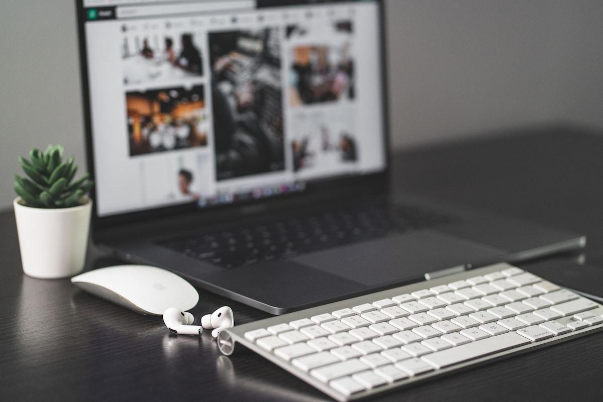 Pessoa escolhendo foto em notebook em banco de imagens trabalhando sobre mesa cinzenta de madeira com teclado, mouse e fone de ouvido sem fio ao redor do mesmo, todos na cor branca, além de vaso branco pequeno com planta de interiores
