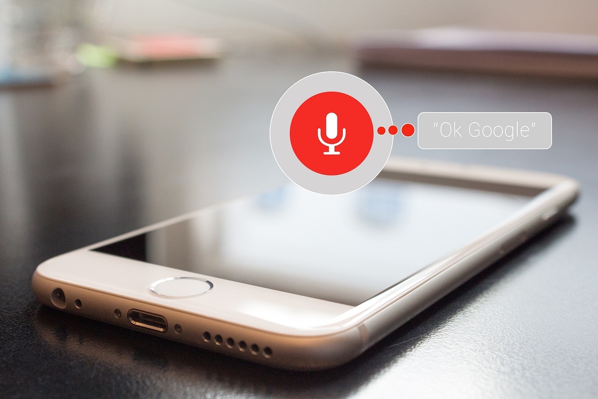 Answer Engine Optimization representado por smartphone sobre mesa escura com símbolo de microfone e frase "Ok Google", uma das formas mais comuns dos usuários fazerem suas pesquisas em formato de perguntas aos buscadores