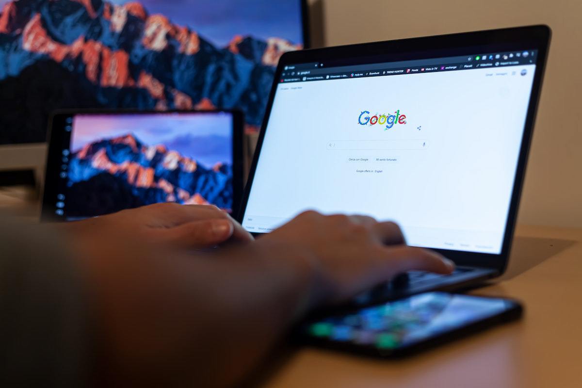 Notebook: Anunciar no Google Pessoa acessando Google em notebook com smartphone do lado, além de tablet com monitor ao fundo da imagem em desfoco ao fundo