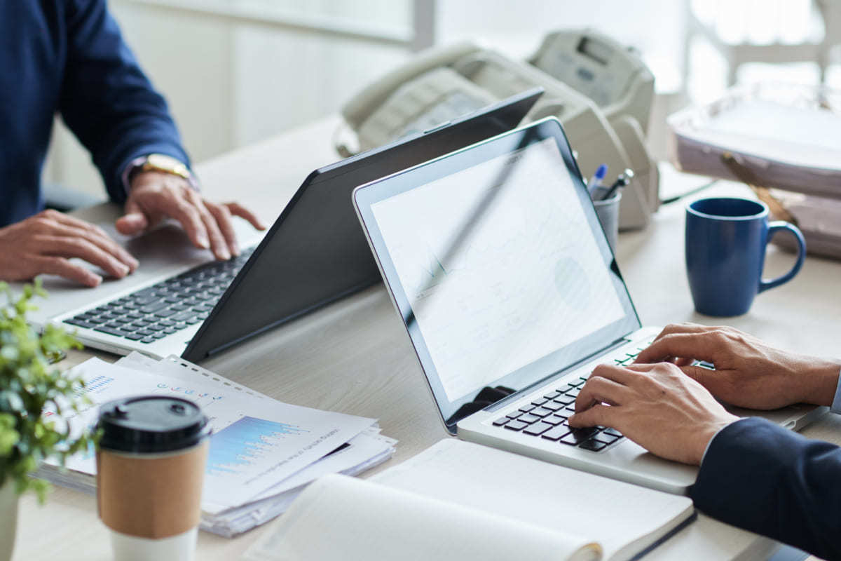 Escritório: Atendimento omnichannel e-commerce Dois homens sentados frente à frente usando notebooks sobre mesa branca com itens de escritório e canecas de café, bem como copo descartável para essa bebida, realizando Atendimento omnichannel e-commerce