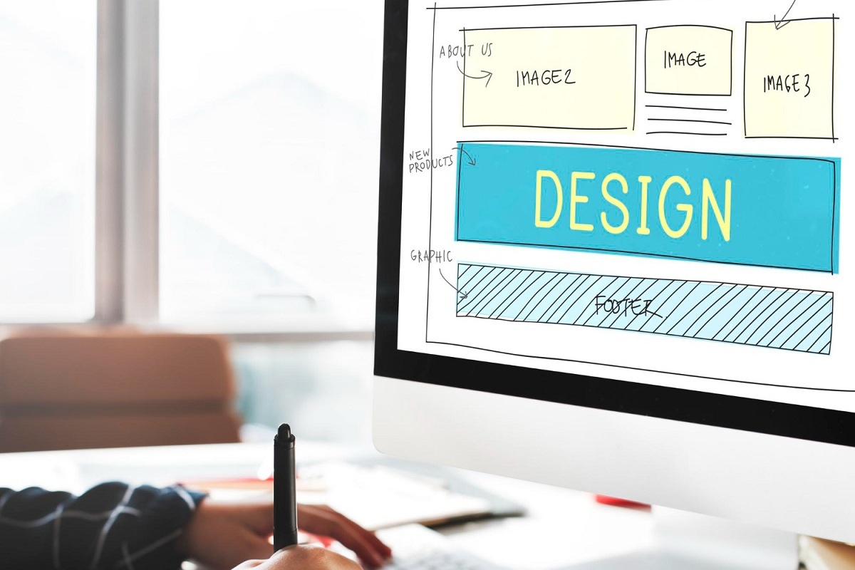 Pessoa fazendo projeto de layout ou design de site em computador com grande monitor sobre mesa e diante de janelas grandes de vidro de edifício enquanto anota algo em uma agenda com caneta preta