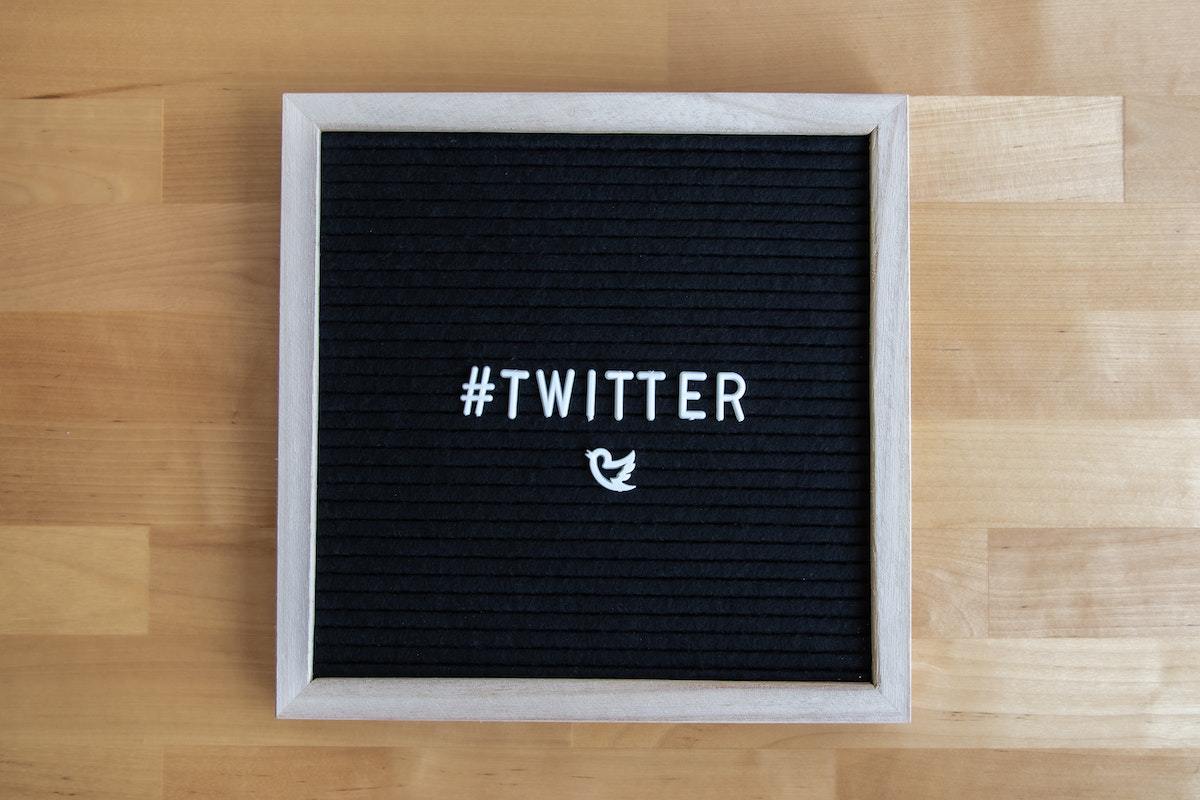 Letreiro preto com nome da rede social Twitter em branco sobre mesa de madeira, além de conter símbolo do mesmo e hashtag