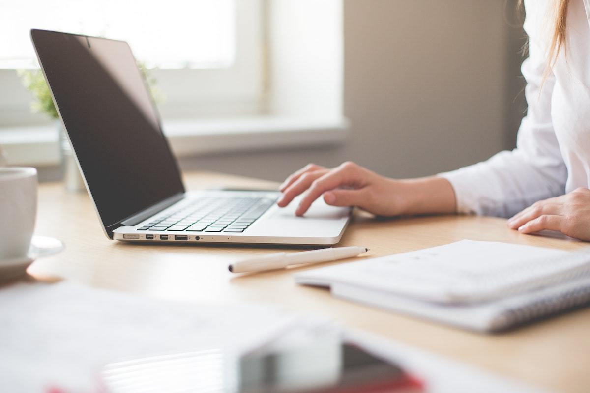 Mulher trabalhando estratégia de Customer insight em escritório usando notebook sobre mesa grande de madeira, com papéis e caderno, além de lápis, ao seu redor e espalhados sobre a mesma mesa