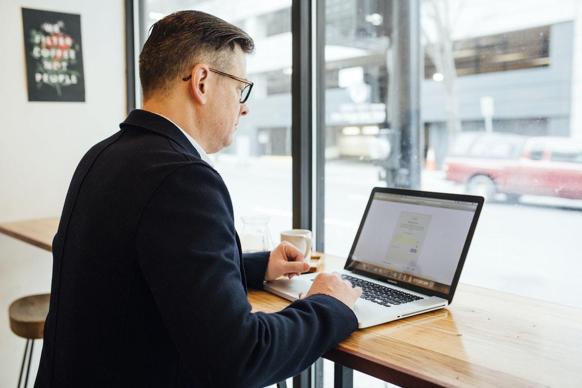Homem vestindo terno usando notebook para analisar estratégia de Customer insight em mesa de madeira de local que parece cafeteria ou restaurante de frente à janela de vidro grande que lhe dá visão da rua, onde passa um carro vermelho, em desfoco na foto