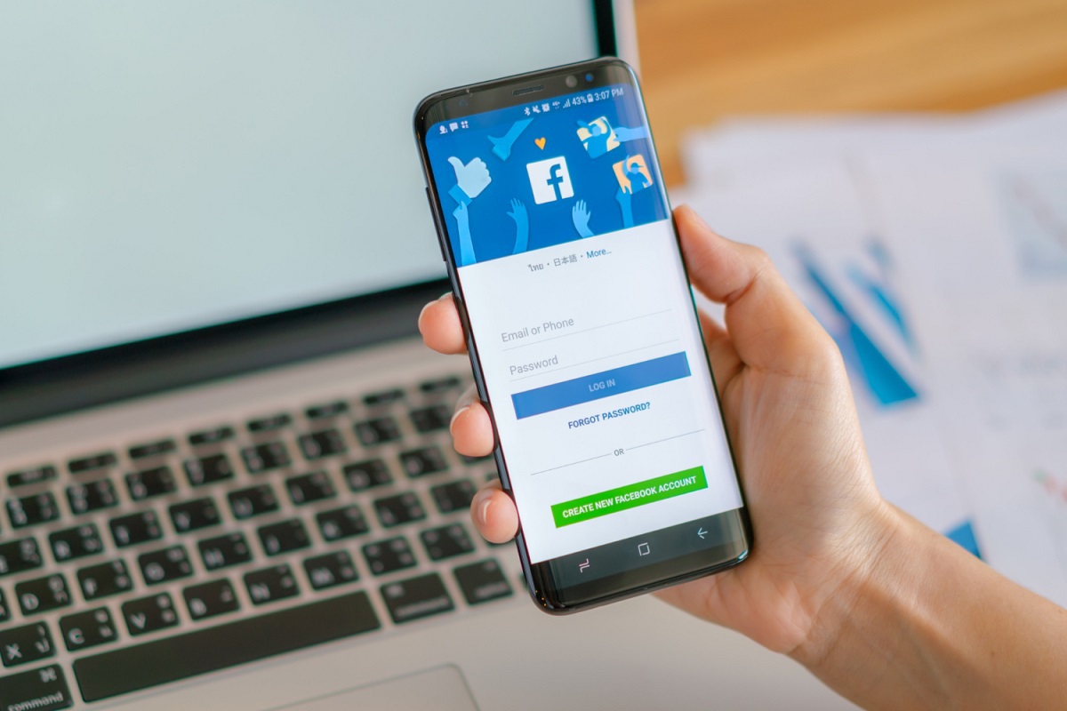 Pessoa segurando celular com app do Facebook aberto em tela de login diante de mesa com notebook e papéis com dados impressos