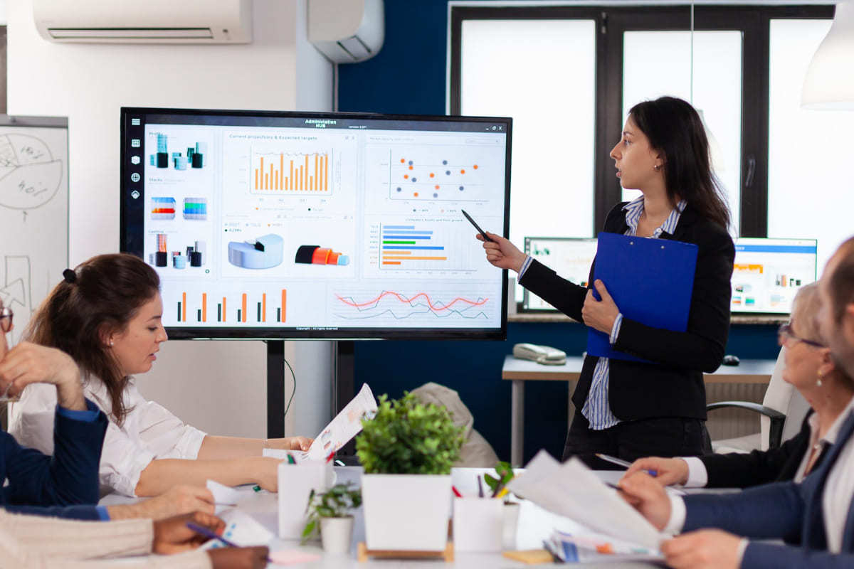 Mulher fazendo Demonstração de produto em reunião com colegas apontando dados em monitor grande