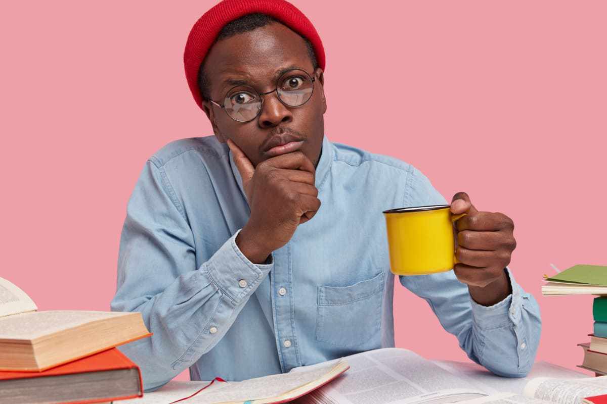 Homem com caneca amarela na mão com ar de confuso com livros e revistas sobre mesa enquanto faz estudos