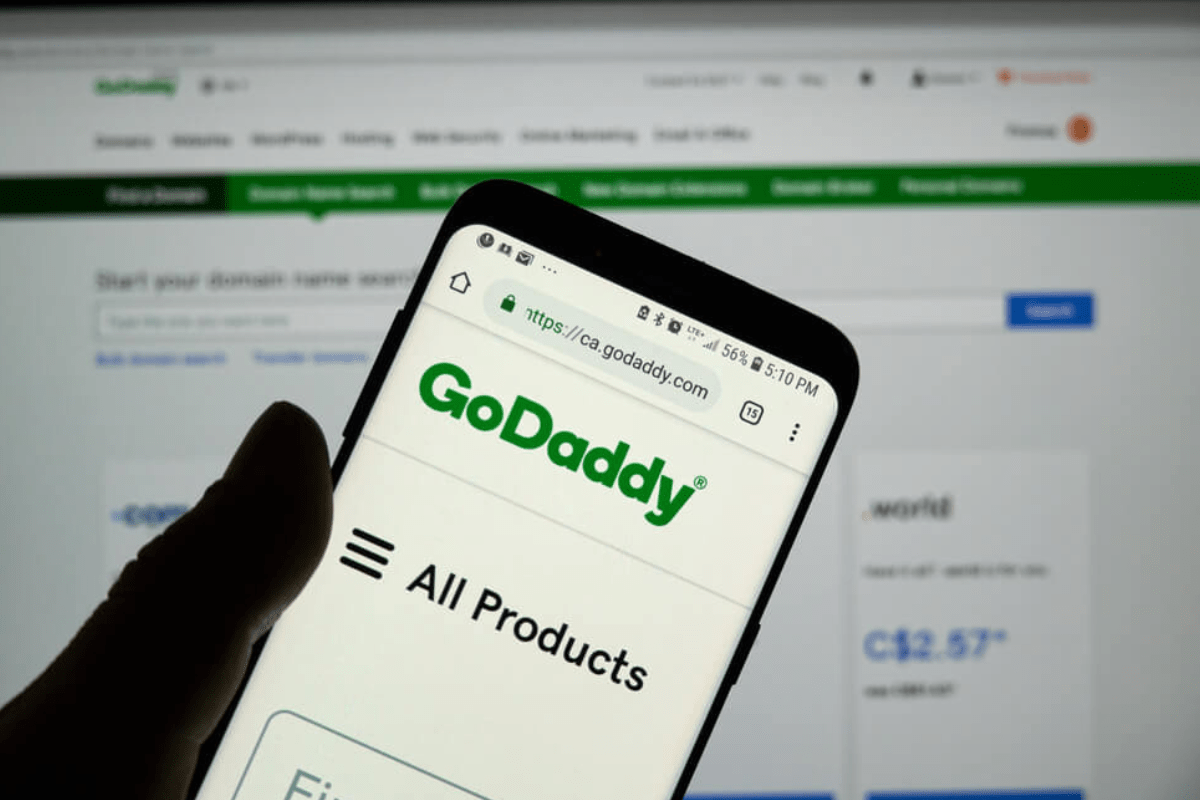 Celular: Go Daddy Pessoa segurando celular com mão esquerda acessando a Go Daddy, diante de tela de computador, também usando a solução