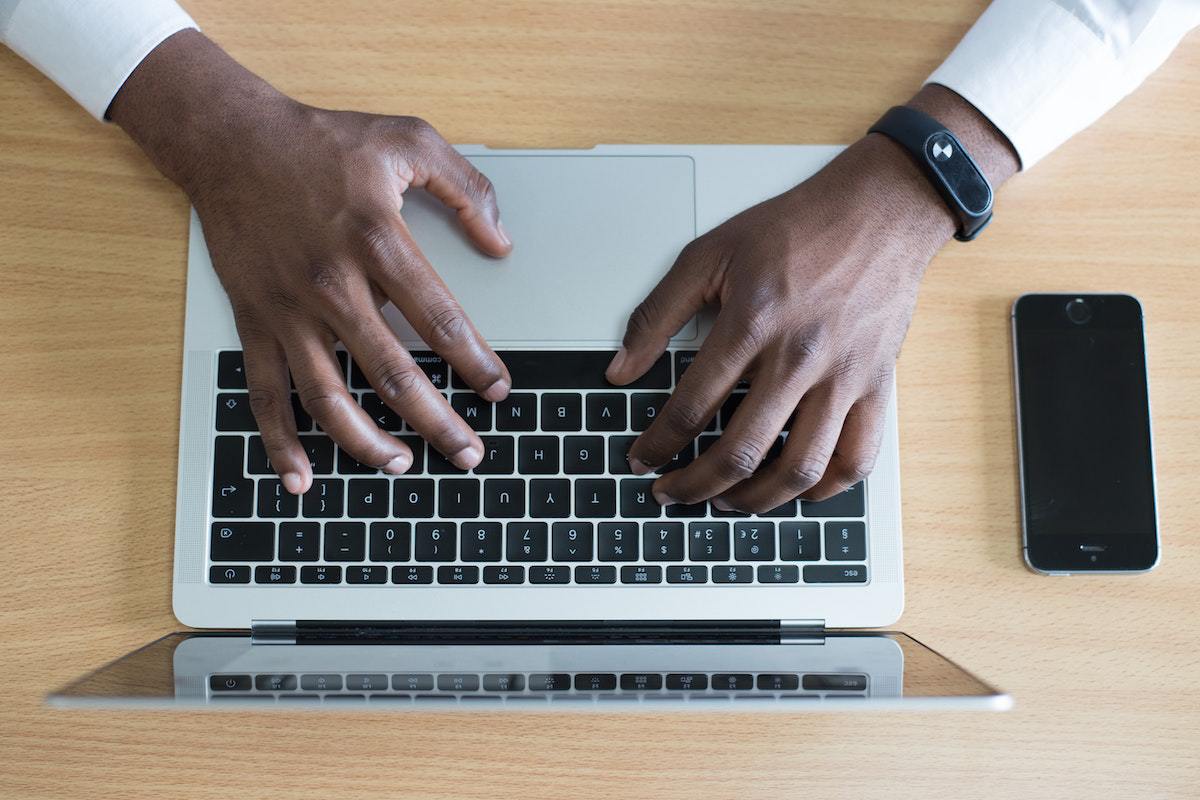 Homem usando notebook com mãos sobre o seu teclado enquanto navega na internet por meio do aparelho sobre mesa de madeira com smartphone preto à sua esquerda