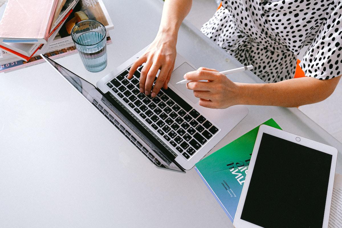Pessoa fazendo seu trabalho em Home office e produtividade a mil com notebook e tablet ao seu lado, além de livros e agenda com copo de água cheio feito em vidro na cor azul à direita do trabalhador