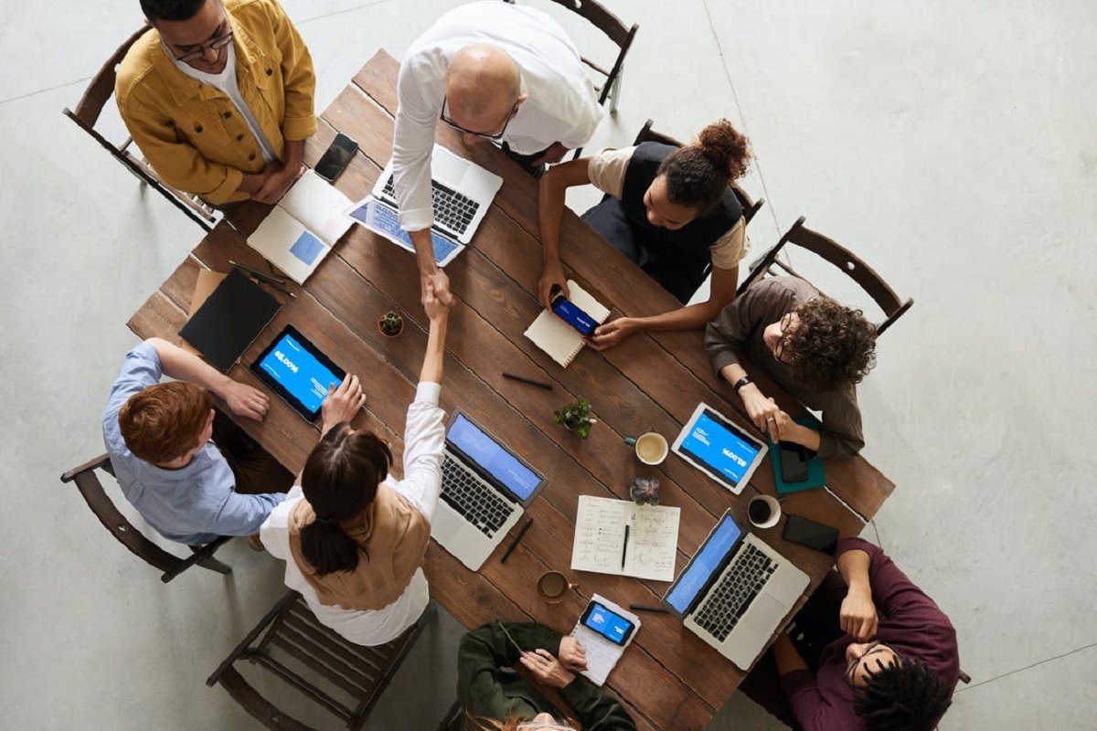 Ideias de empreendedorismo debatidas em reunião ao redor de mesa de madeira em empresa enquanto pessoas usam notebooks e tablets em foto tirada de cima