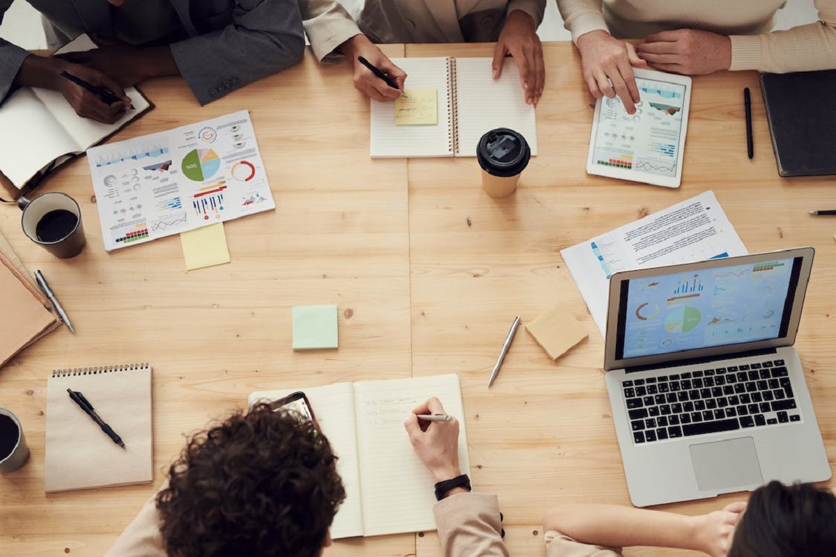 Pessoas em local de trabalho ao redor de mesa de madeira usando tablets e notebooks e falando sobre Ideias de empreendedorismo