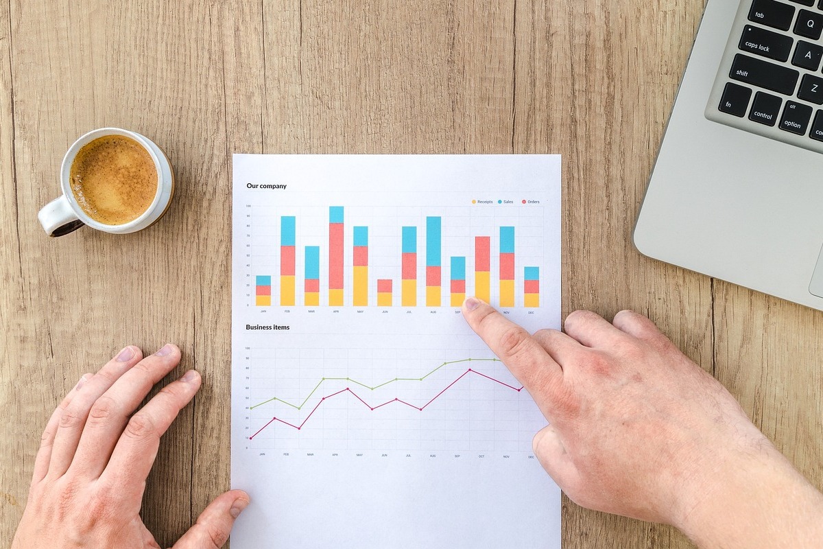 Gráficos: Kissmetrics Pessoa analisando gráficos impressos em folha a4 extraídos do Kissmetrics sobre mesa de madeira e ao lado de notebook e xícara de café espresso