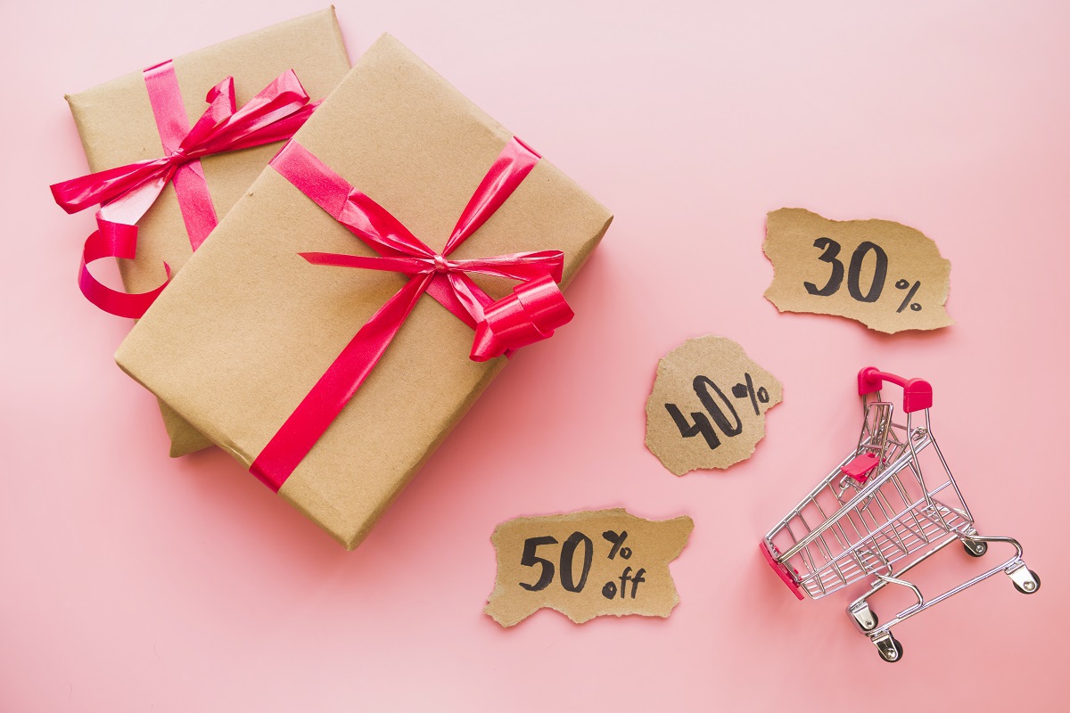 Descontos: Live marketing Caixas de presente com laço rosa sobre superfície rosa ao lado de balões com porcentagens que representam descontos e com carrinho de compras pequeno em montagem para Live marketing