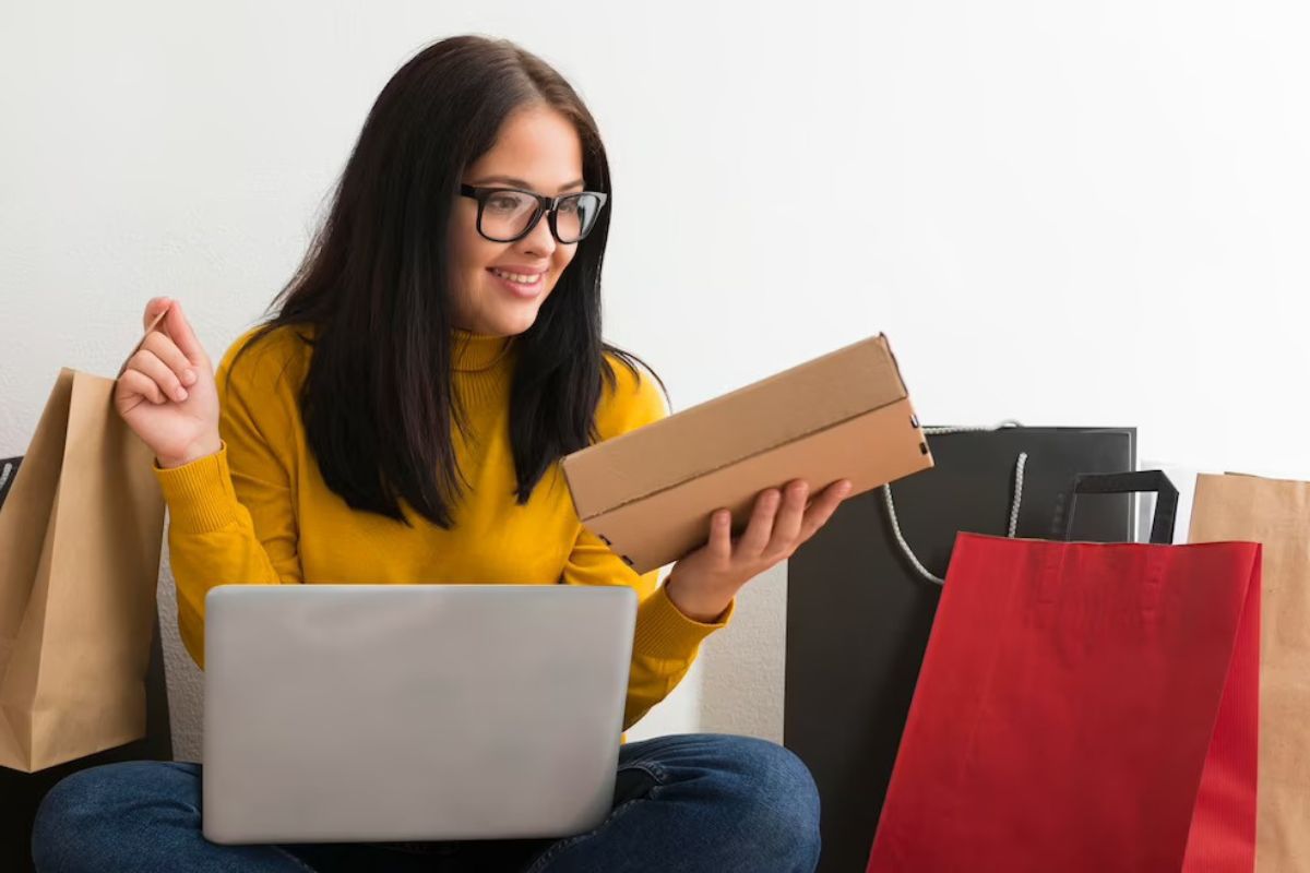 Mulher sentada em sofá com sacolas de compras, além de caixa, bem como notebook em seu colo, representando tema Marketing convencional