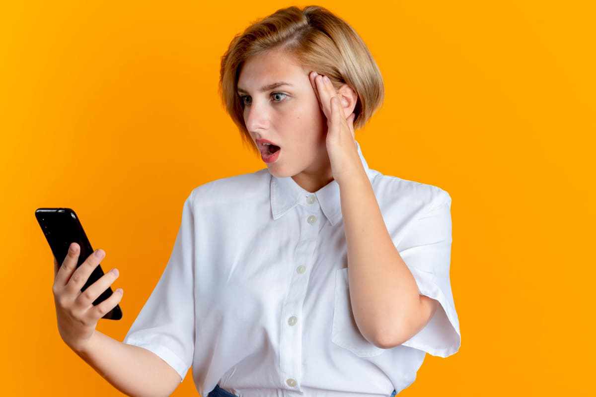 Mulher com ar de surpresa e feição que mostra esse sentimento com mão na lateral do rosto e cabelo, enquanto olha algo em celular, que provocou tal reação na mesma com Metodologia AIDA, diante de parede laranja ao fundo da imagem ou foto