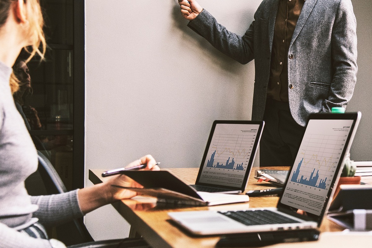 Escritório: Métricas de negócios Mulher em escritório com notebooks analisando gráficos e Métricas de negócios com homem diante da mesma usando paletó enquanto parece perguntar algo à mesma