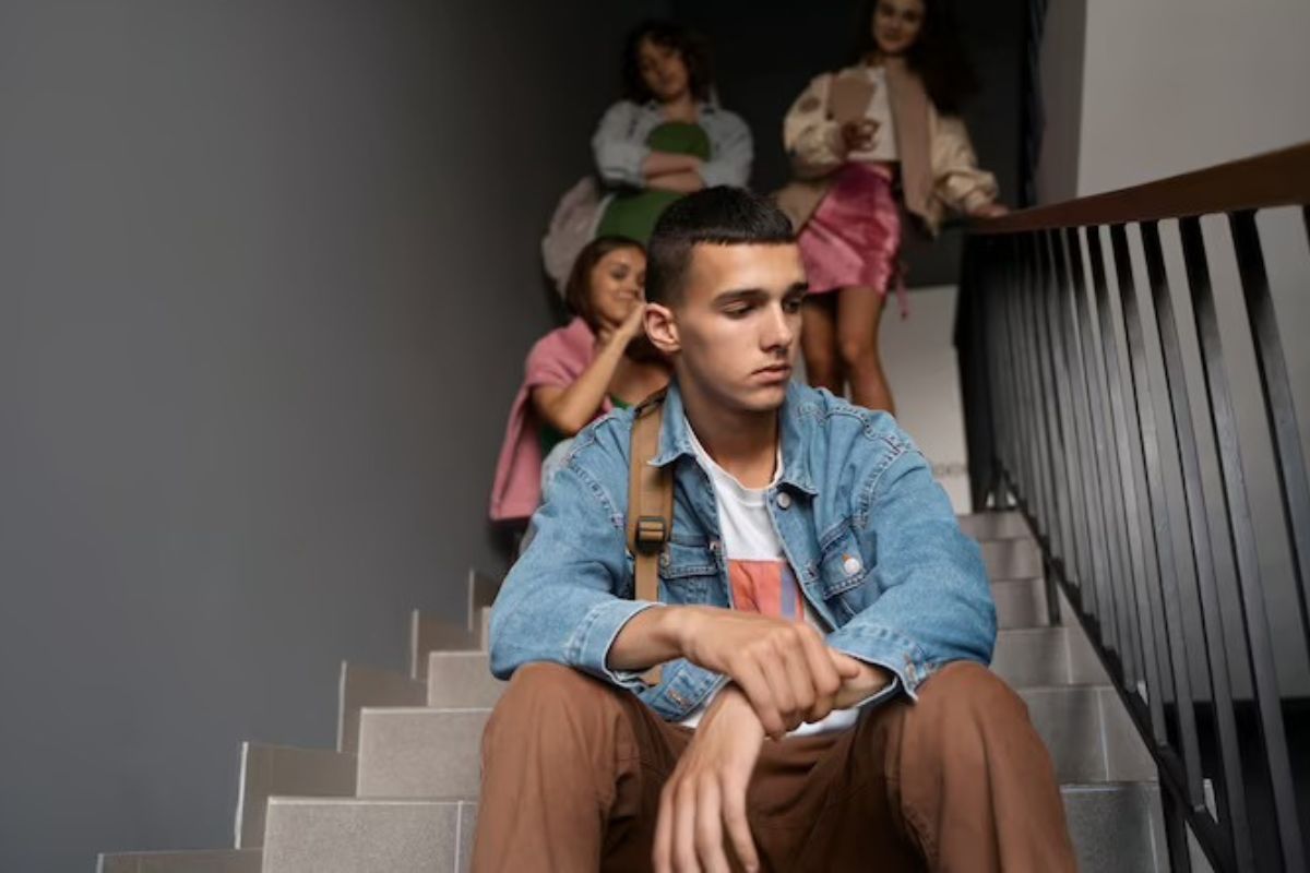 Jovem distraído com seu pensamento sentado em escada com pessoas reunidas atrás do mesmo conversando sorridentes