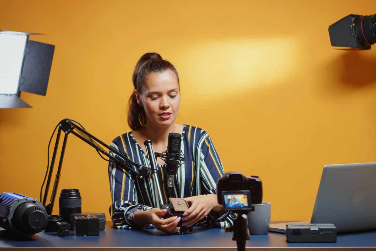 Mulher diante de parede laranja sentada na frente de bancada com microfone de poscast articulado gravando passo a passo para Montar uma loja virtual
