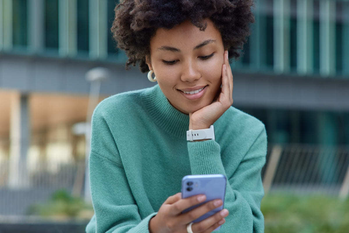 Celular: Nicho para blog Mulher sentada em local que parece ser calçada com prédio em desfoco à suas costas e mão no queixo sorridente usando seu celular