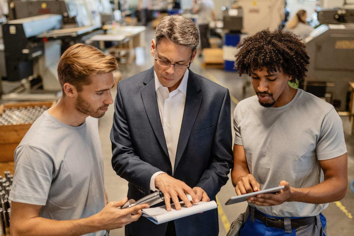 Líder: POP Líder em interior de empresa mostrando procedimento POP para dois colaboradores, um em cada lado seu, de pé, analisando folhas de papel
