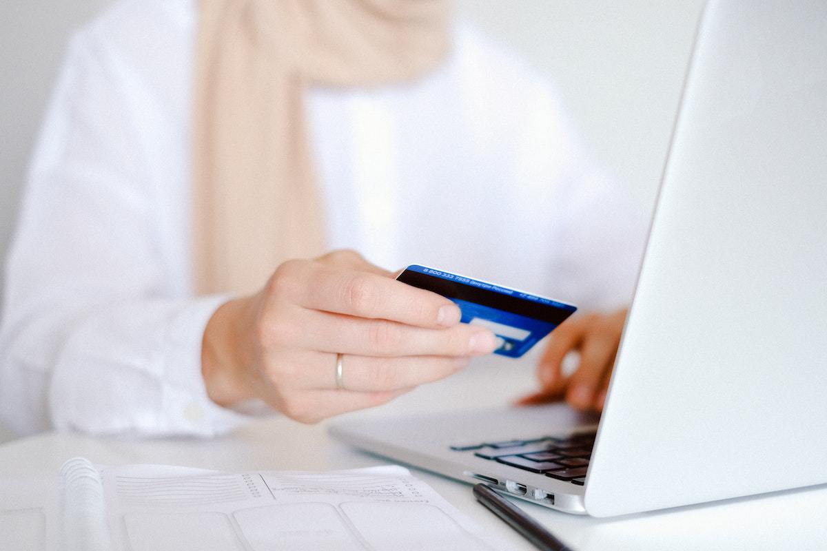 Pessoa com cartão de crédito nas mãos e sentada diante de mesa branca usando notebook para fazer Pagamentos online