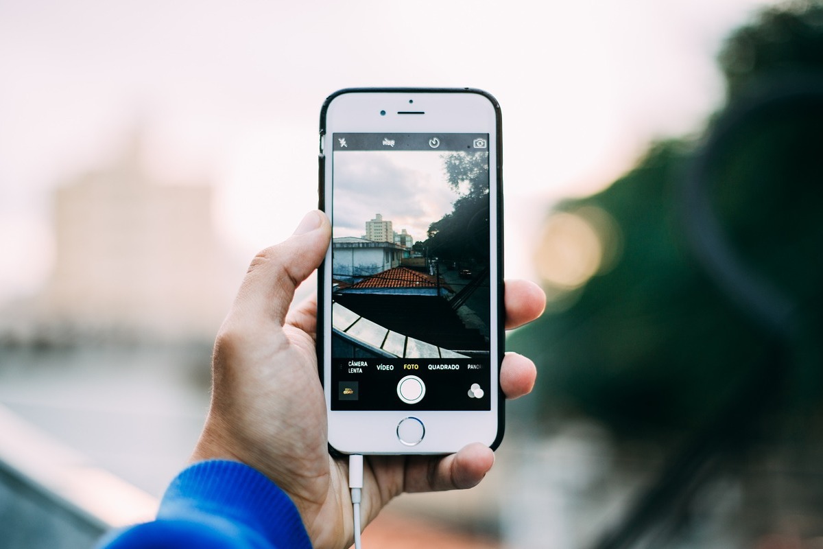 Pessoa com smartphone branco com case preto nas mãos com cabo de, possivelmente, fone de ouvido com câmera aberta apontando para um local
