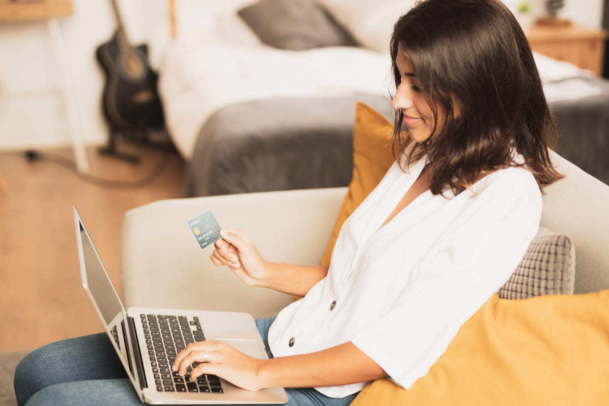 Cartão: Psicologia do consumo no e-commerce Mulher sentada em sofá de casa segurando notebook e cartão de crédito em representação à Psicologia do consumo no e-commerce