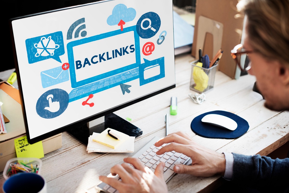 Homem trabalhando em computador sobre mesa rústica de madeira com tela grande do monitor exibindo montagem com símbolos web espalhados na imagem e título BACKLINKS, que é uma das estratégias que podem ser usadas no SEO para landing pages, bem centralizado, com itens de escritório próximos ao mesmo espalhados ao seu redor