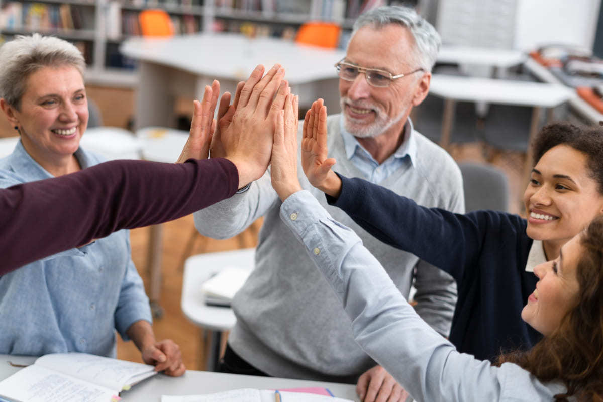Equipe: Sistema gestão educacional Equipe com homem e mulheres em ambiente que parece interior de biblioteca tocam as palmas das mãos em comemoração