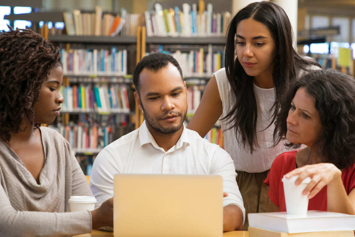Homem e três mulheres sentados diante de mesa usando Sistema gestão educacional em notebook com prateleira de livros ao fundo