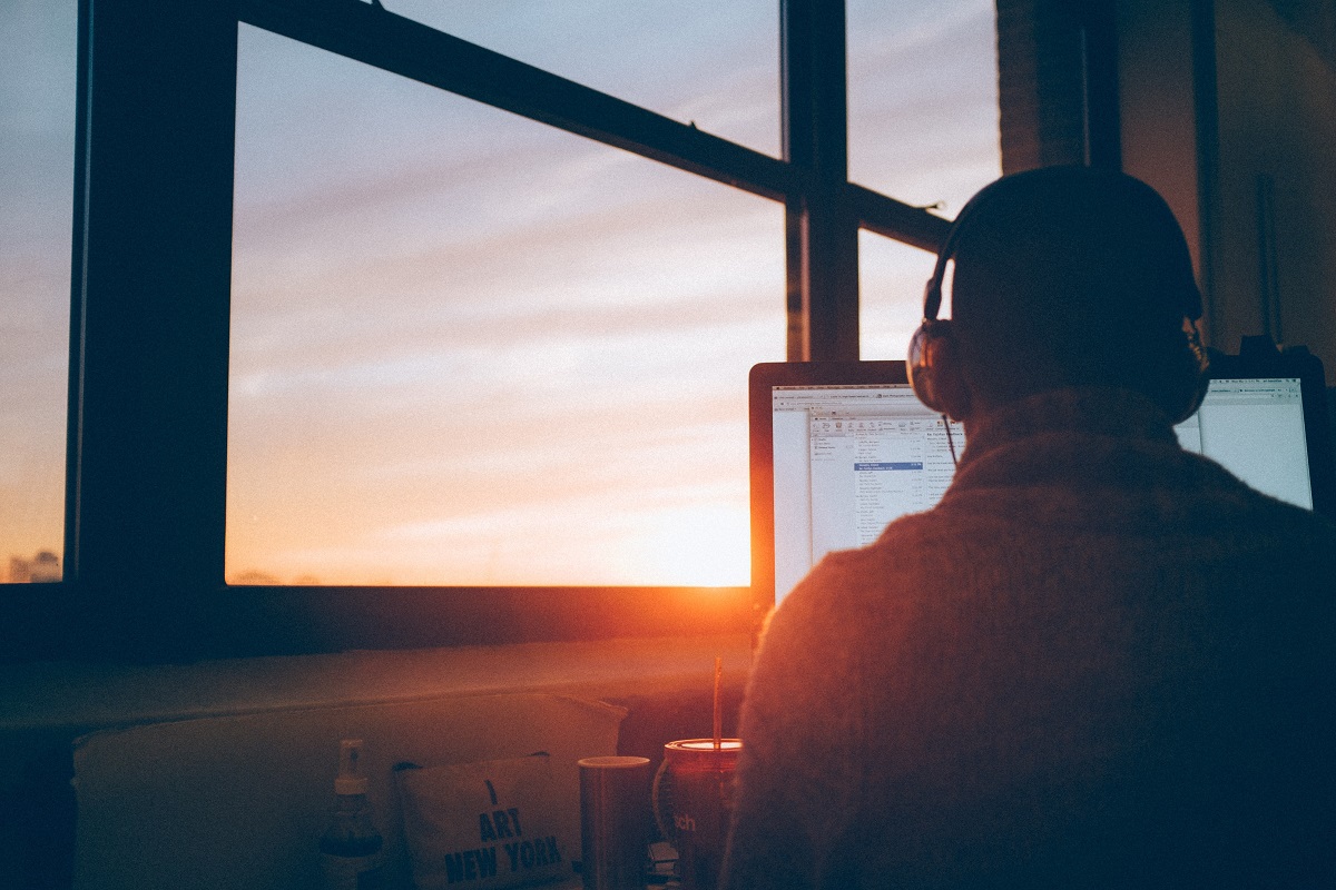 Homem de costas trabalhando diante de computador com fundo da foto mostrando um bonito por do sol que ele vê através de uma janela de vidro do edifício onde está
