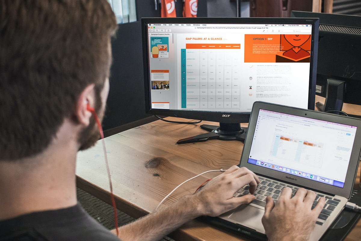 Profissional durante processo de criação de arte tipo Web design, usando programas em laptop conectado à monitor grande à sua esquerda e ao fundo, tudo sobre mesa de madeira, enquanto o indivíduo usa fone de ouvido com fio na cor vermelha
