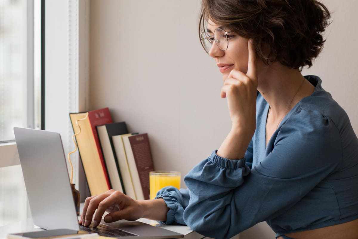 Notebook: XML Mulher com mão no queixo usando notebook ao lado de livros escorados na parede diante de janela de vidro de apartamento