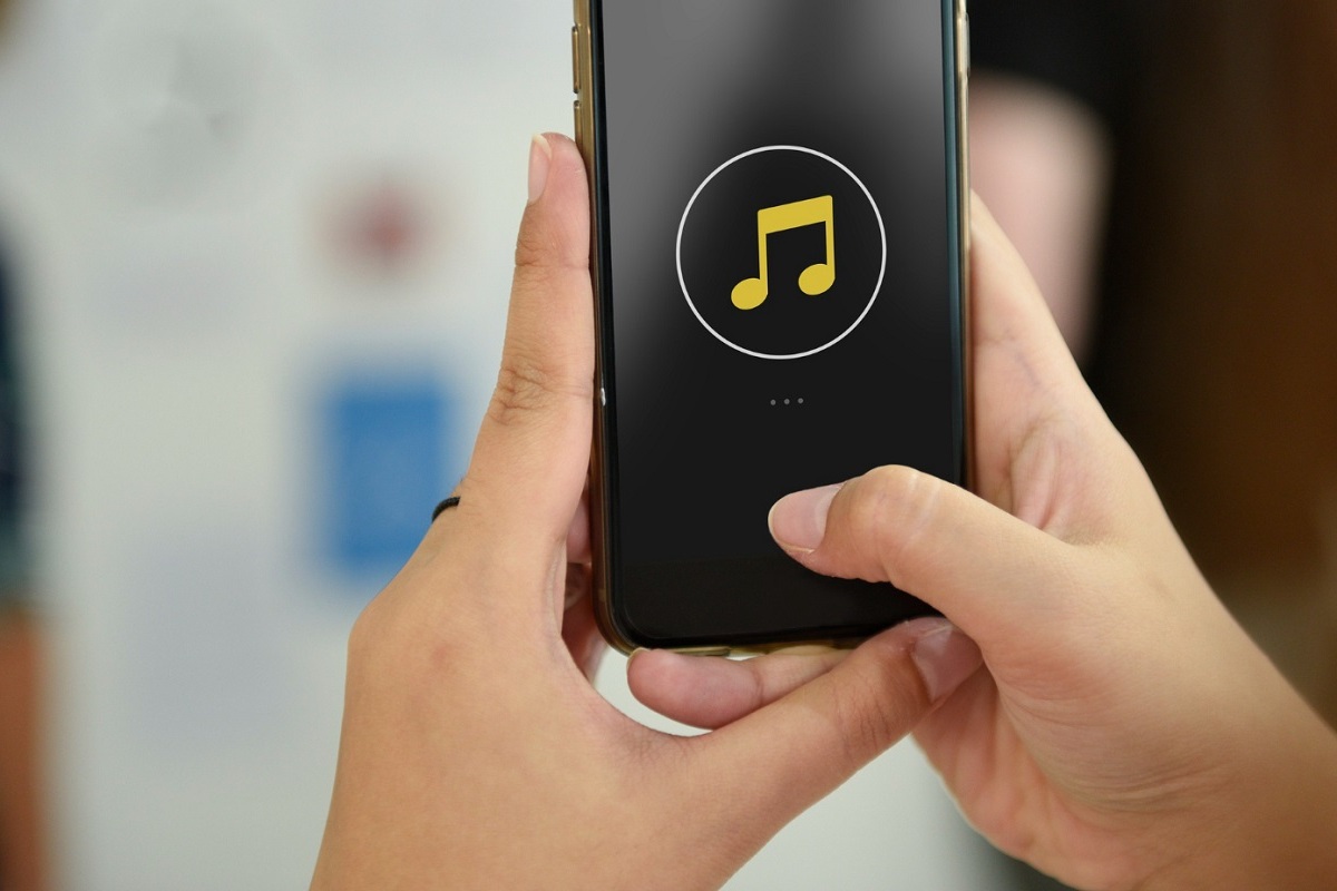 Usuário segurando celular com sua tela exibindo símbolo de música ou nota musical diante de tela de outro dispositivo, similar à monitor, em desfoco ao fundo da imagem