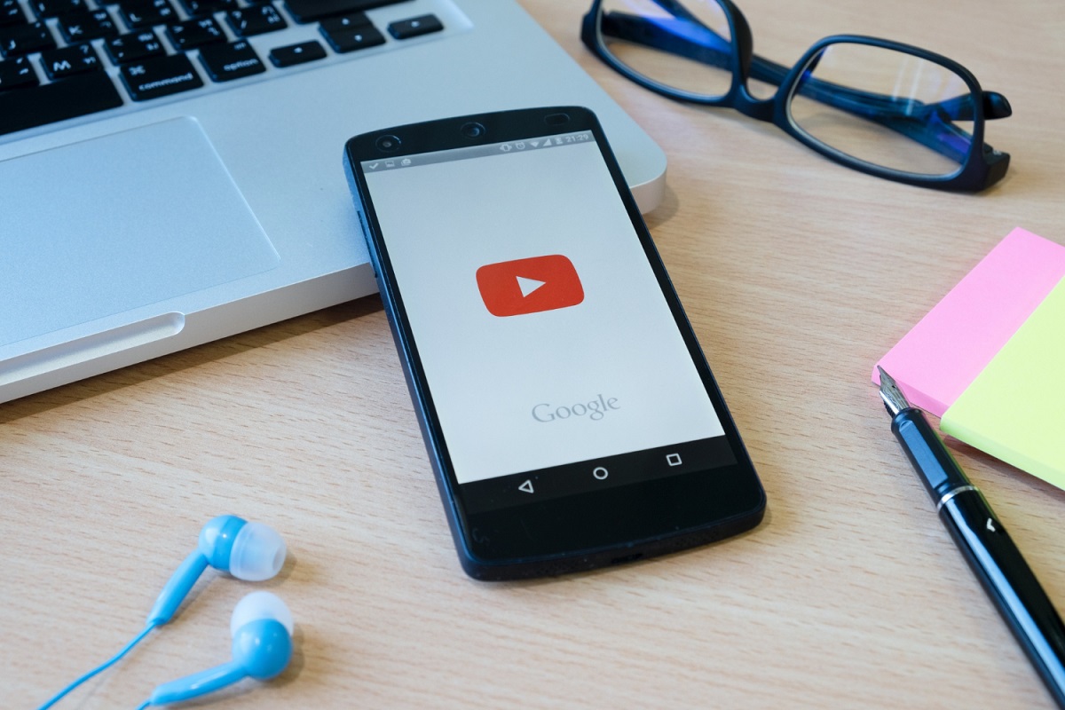 App: YouTube Music Celular sobre mesa de madeira apoiado em teclado de notebook enquanto inicia app do YouTube Music